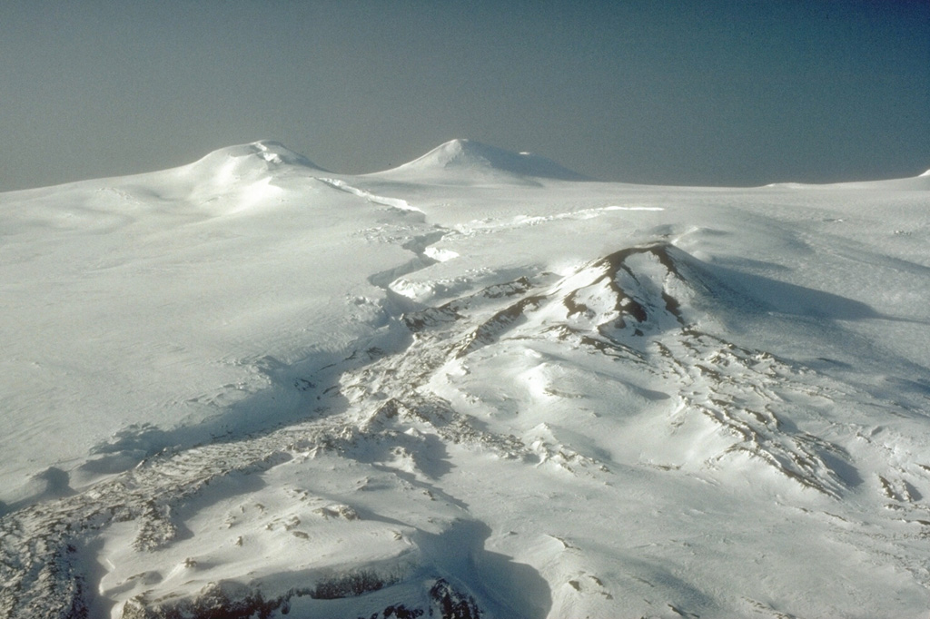 The summit area of Westdahl, located on the southwestern part of Unimak Island in the eastern Aleutians, is seen here from the east. Westdahl Peak (left) and Faris Peak (right) formed on the broad main edifice. The scoria cone in the center was the principal eruption site for the 1991-92 activity, producing the snow-covered lava flow in the foreground. . The sinuous fissure cutting across the glacial icecap from the summit formed in the opening phases of the eruption and produced lava fountaining. Photo by C.F. Zeillemaker, 1993 (U.S. Fish and Wildlife Service, courtesy of Alaska Volcano Observatory).