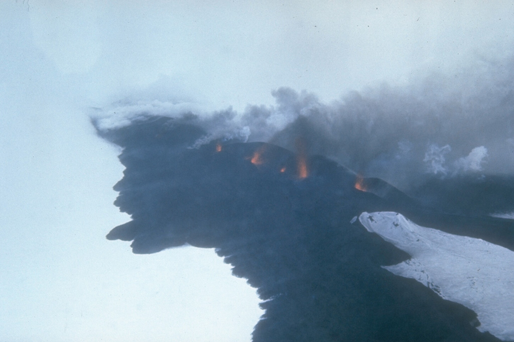 Lava fountains rise above a fissure on Seguam volcano on 8 March 1977. This view from the south shows part of the 1.5-km-long eruptive fissure SE of Pyre Peak. Black lava flows move across the snow from the fissure and diverge around an older cone to the lower right. Pyre Peak, within the westernmost of two calderas, is surrounded by fresh lava fields. Photo by U.S. Coast Guard, 1977 (courtesy of Alaska Volcano Observatory).