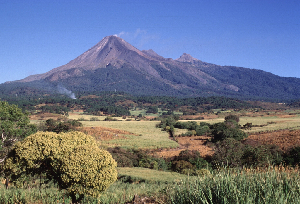Colima (left), with a diffuse gas plume drifting from its summit, and Nevado de Colima (right) form a pair of edifices comprising the Colima volcanic complex. The modern Colima cone was constructed during the past 2,600 years within a large horseshoe-shaped flank collapse scarp that opens towards the south. The modern edifice has overtopped the eastern caldera rim (roughly corresponding to the vegetation line), and unvegetated lava flows that were emplaced during 1975-76 are visible down the outer flanks. Photo by Lee Siebert, 1997 (Smithsonian Institution).