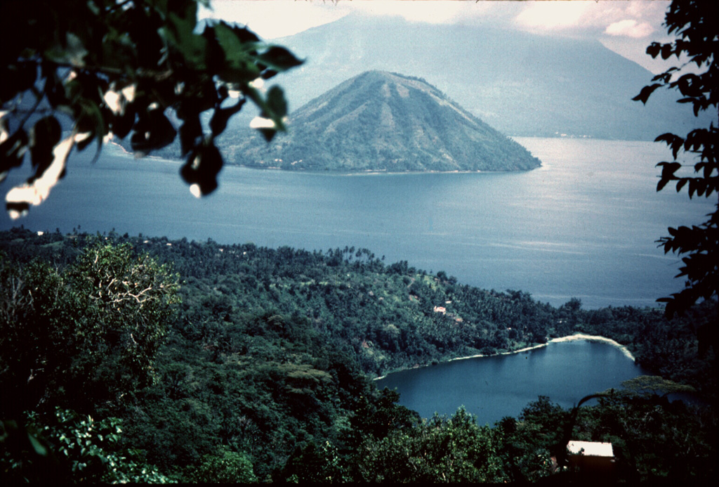 The small conical island of Maitara is between Ternate and Tidore in northern Maluku off the W coast of Halmahera. In the background, its summit hidden in the clouds over southern Tidore Island, is symmetrical Kie Matabu volcano.  Telaga volcano, a truncated caldera at the NE end of Tidore Island, is obscured behind the leaves at the upper left.  The lake-filled crater in the foreground is Ngade maar on the southern flank of Gamalama volcano. Photo by Jack Lockwood, 1980 (U.S. Geological Survey).