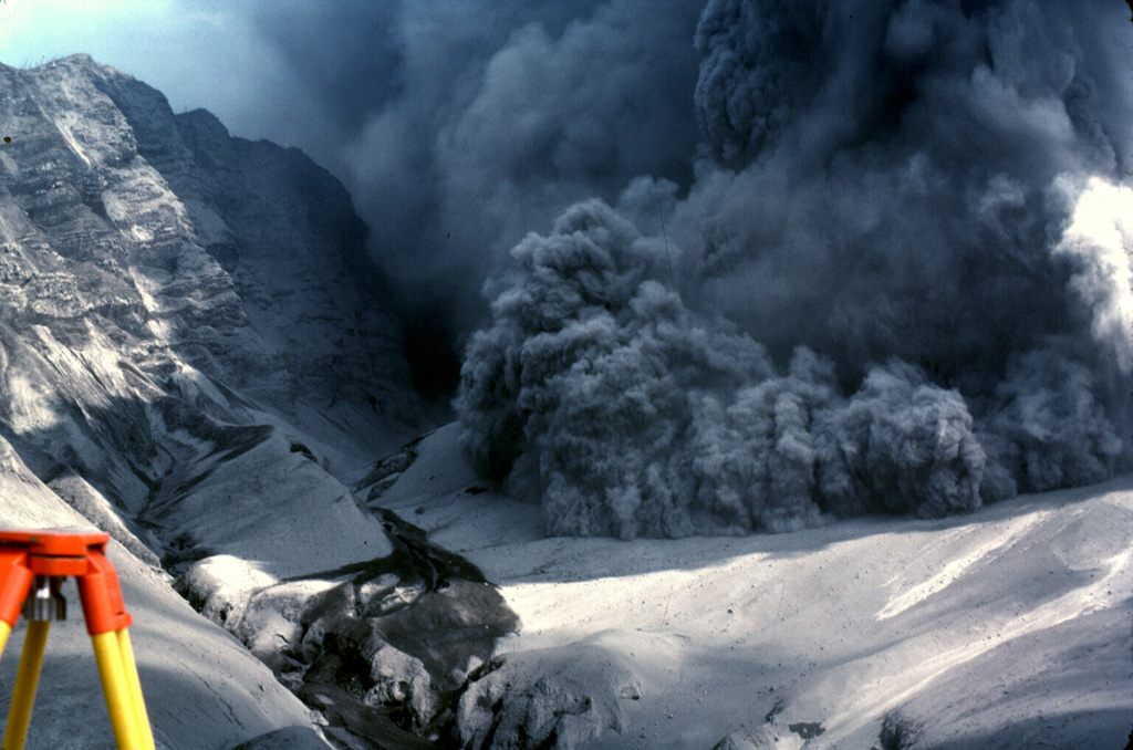 An explosive eruption from Galunggung on 7 August 1982 is accompanied by a pyroclastic flow advancing over the crater rim towards a reflector station that was being measured using the tripod to the lower left. The photo was taken from Butik Pasir Bentag, about 2 km from the crater. The 1-km-high ash-covered cliff to the left is a wall of the breached caldera. Photo by Jack Lockwood, 1982 (U.S. Geological Survey).