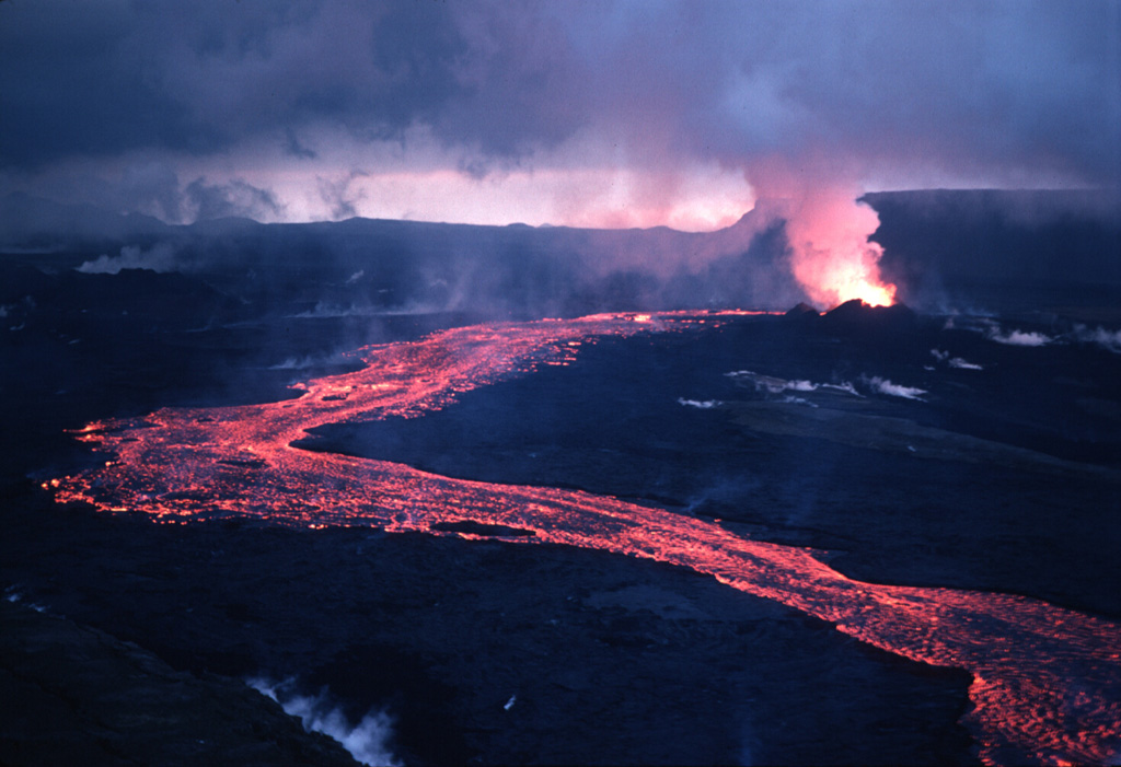 An incandescent lava flow travels downslope from the vent at the upper right during a September 1984 eruption from Krafla volcano. The eruption initially began on 4 September along an 8.5-km-long fissure; during later stages activity was localized along singular vents such as the one seen in this 15 September photo. A plume of gas is concentrated over the vent, but steam is also seen rising across much of this photograph, from recent lava flows. Photo by Michael Ryan, 1984 (U.S. Geological Survey).