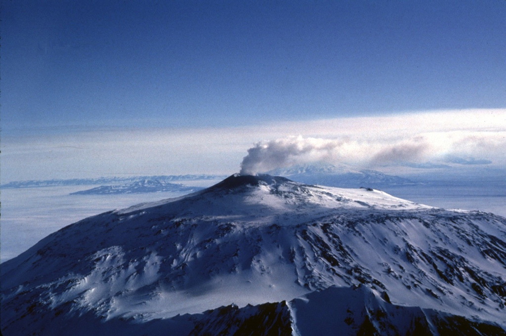 A gas-and-steam plume rises above the Mount Erebus summit crater in 1983. Fang Ridge is in the foreground of this view from the NE. The summit plateau across the center of the photo is the rim of a caldera containing the active summit cone. Photo by Bill Rose, 1983 (Michigan Technological University).