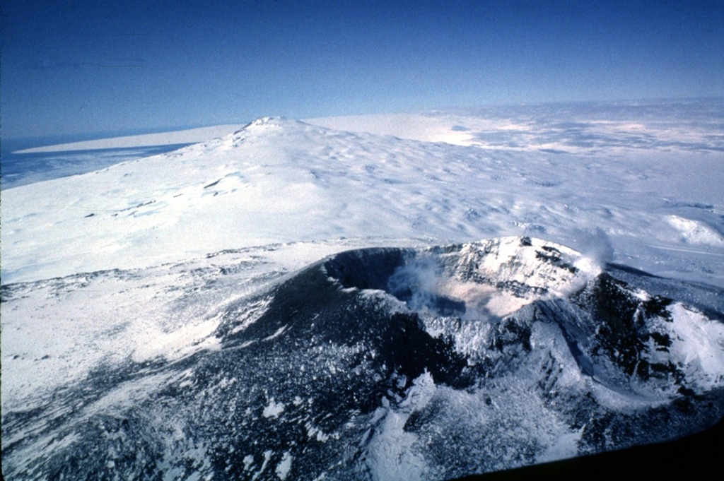Two of the three volcanoes forming Ross Island are seen in this aerial view from the west. The Erebus summit crater that contains an active lava lake is in the foreground, with glaciated Mount Terror in the distance. Numerous scoria cones and lava domes have formed across the flanks. Photo by Bill Rose, 1983 (Michigan Technological University).