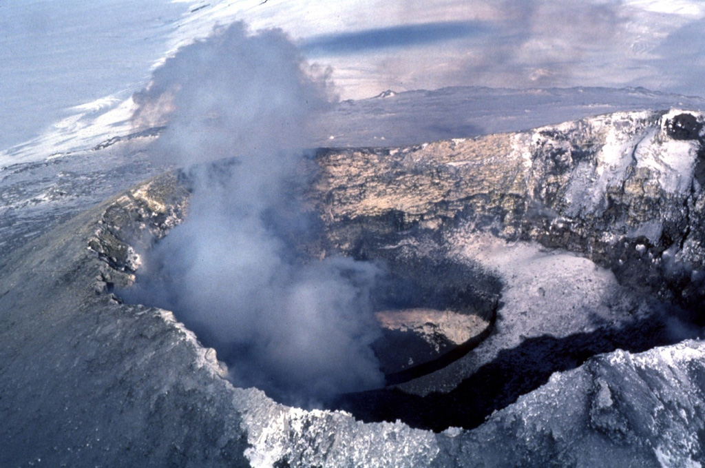 A gas plume rises above the active lava lake at the summit of Erebus in December 1983, which was first observed on 24 December 1972. Lava lake activity and intermittent Strombolian eruptions have been observed during annual scientific expeditions since then. The lava is located at the bottom of an inner 100-m-deep crater, whose snow-covered rim forms a bench at the right (SW) side of the Main Crater in this 1983 photo. Photo by Bill Rose, 1983 (Michigan Technological University).
