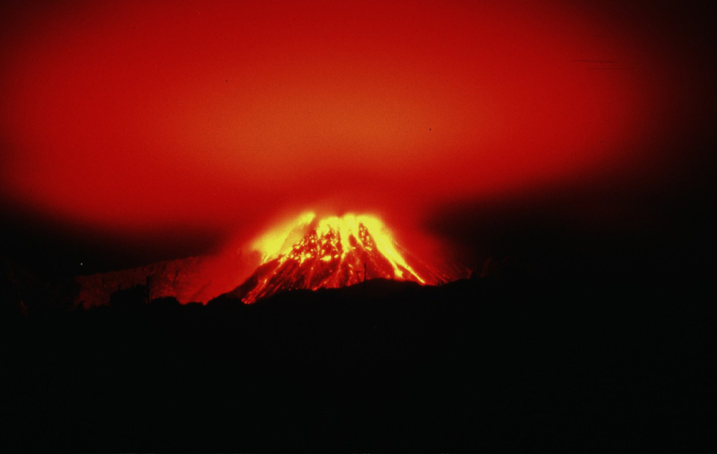 The incandescent lava dome of Soufrière Hills is reflected off the smoke-and-ash column above the volcano on the night of December 26, 1996.  The dome is seen here from Long Ground/White's House on the NE flank, 2 km from the summit.  This 40-second time exposure captures the paths of incandescent rockfalls and block-and-ash flows during a period characterized by one of the highest magma extrusion rates of the eruption. Photo by Mark Davies, 1997 (Montserrat Volcano Observatory).