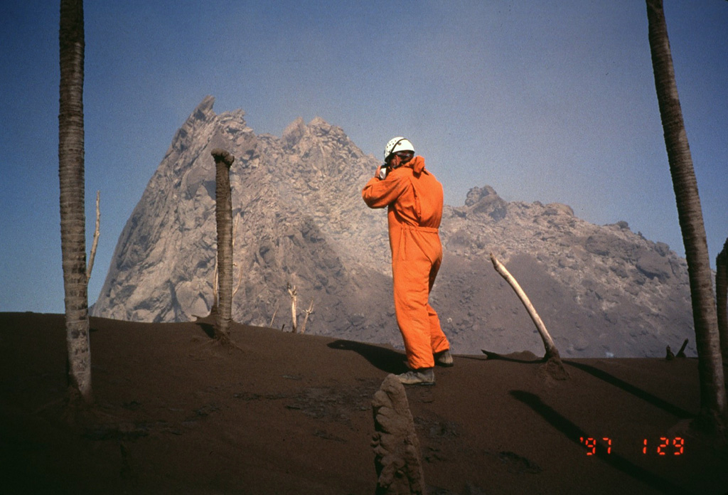 Volcanologist Richard Heard of the Montserrat Volcano Observatory observes the new lava dome in February 1997.  The surface temperature of the lava dome at this time was 650-800 degrees centigrade.  By this time the new dome had grown to a height above that of Chances Peak, the former summit of the volcano and the location of this photo.  A steep-sided spine forms the northern side of the lava dome.  Periodic spine extrusion and subsequent collapse occurred frequently during the eruption.   Photo by Mark Davies, 1997 (Montserrat Volcano Observatory).