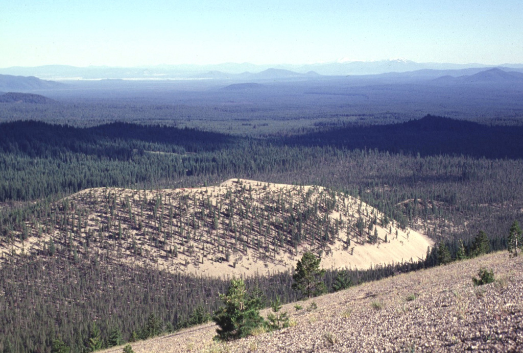 The back side of light-colored Paint Pot Crater and Pumice Stone Mountain, from where the photo was taken, are mantled by white pumice from the eruption that preceded the extrusion of the nearby Little Glass Mountain obsidian flow.  The formation of Paint Pot Crater and the associated extrusion of the lava flow that forms the sparsely vegetated area south (right) of the cone occurred about 1100-1150 years ago, only shortly before the major Little Glass Mountain eruption. Photo by Lee Siebert, 1998 (Smithsonian Institution).