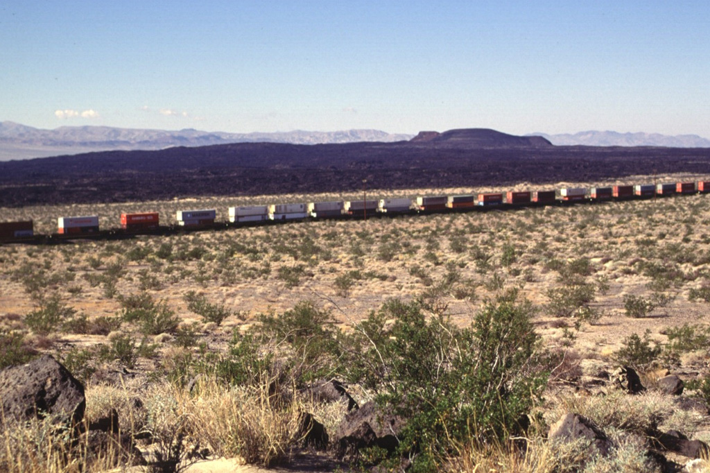 A freight train passes Pisgah Crater and its associated lava field.  The 100-m-high cone is the most prominent feature of a lava field that covers 100 km2.  The irregular shape of the cone results from the mining of aggregate.  The composite lava flows extend up to 8 km to the SE and in a narrow lobe 18 km to the NW.  Most of the flows are pahoehoe, but some of the eastern flows are aa.  Other cones and lava flows of the Lavic Lake volcanic field are located nearby. Photo by Lee Siebert, 1997 (Smithsonian Institution).