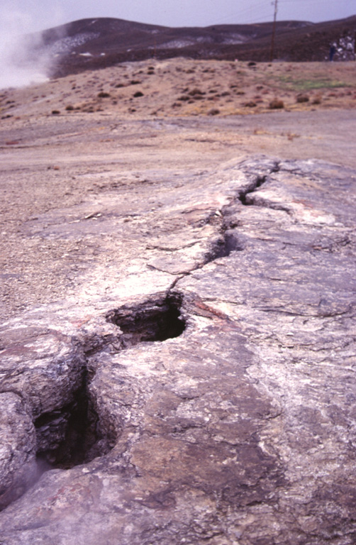 A now-inactive fissure cuts the surface of a sinter mound at Steamboat Springs in Nevada.  Steam from a hot springs rises at the upper left.  Steamboat Springs, an area of active geothermal development, currently displays hot springs, a large number of steam vents and fumaroles structurally related to regional faults.  The area once contained about 20 small geysers, but none are currently active. Photo by Paul Kimberly, 1997 (Smithsonian Institution).