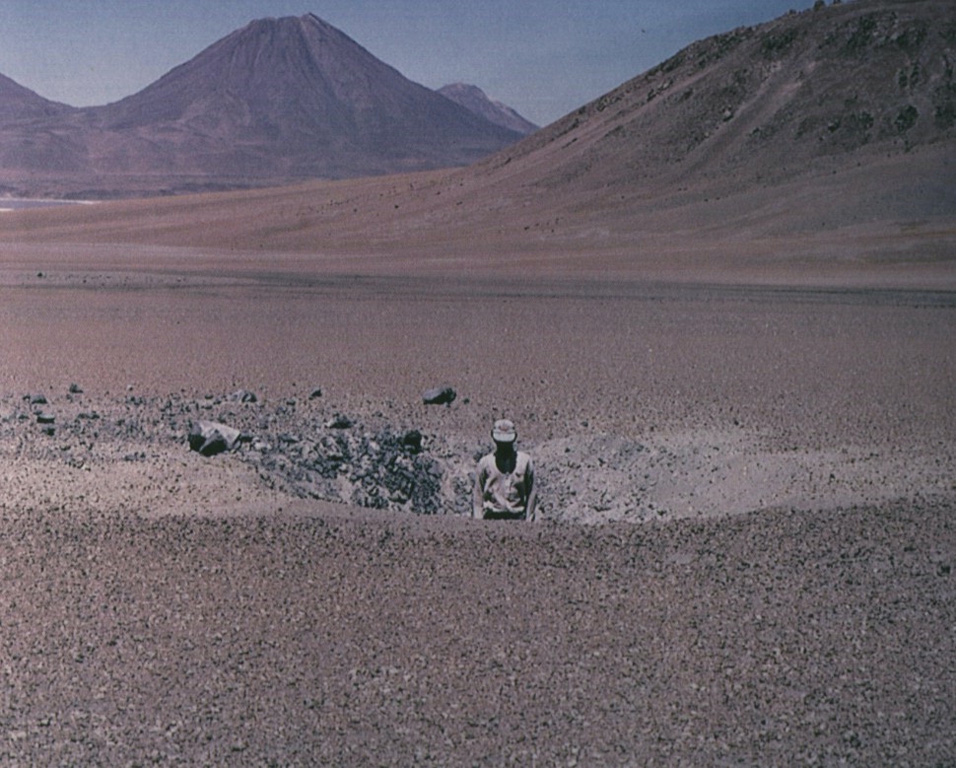 This impact crater, 6 m wide and 1.5 m deep was produced by ejection of a 1.5 cu m ballistic block that traveled 5 km from the summit crater of Láscar (out of view to the right).  An explosive eruption on February 20, 1990 destroyed 10-30% of the summit crater lava dome that had been emplaced in 1989 and produced an 18-km-high ash cloud.  Aguas Calientes volcano rises on the horizon.   Photo by Oscar González-Ferrán, 1990 (University of Chile).