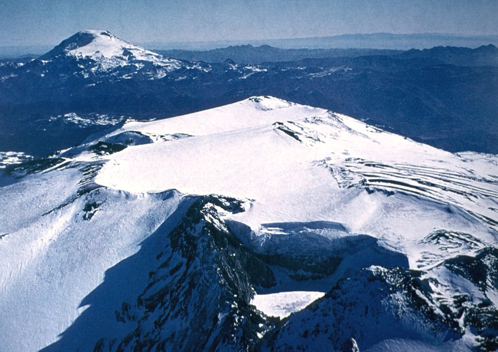 The broad glacier-covered summit of Volcán Copahue along the Chile/Argentina border is seen from the SE with conical Callaqui volcano in the distance.  The lake-filled active crater of Copahue (lower center) has been the site of historical eruptions.  The composite cone was constructed along the Chile/Argentina border within an 8-km-wide caldera formed 0.6 million years ago.  The eastern summit crater is part of a 2-km-long, ENE-WSW line of nine craters that cuts across the western rim of the caldera. Photo by Oscar González-Ferrán, 1992 (University of Chile).