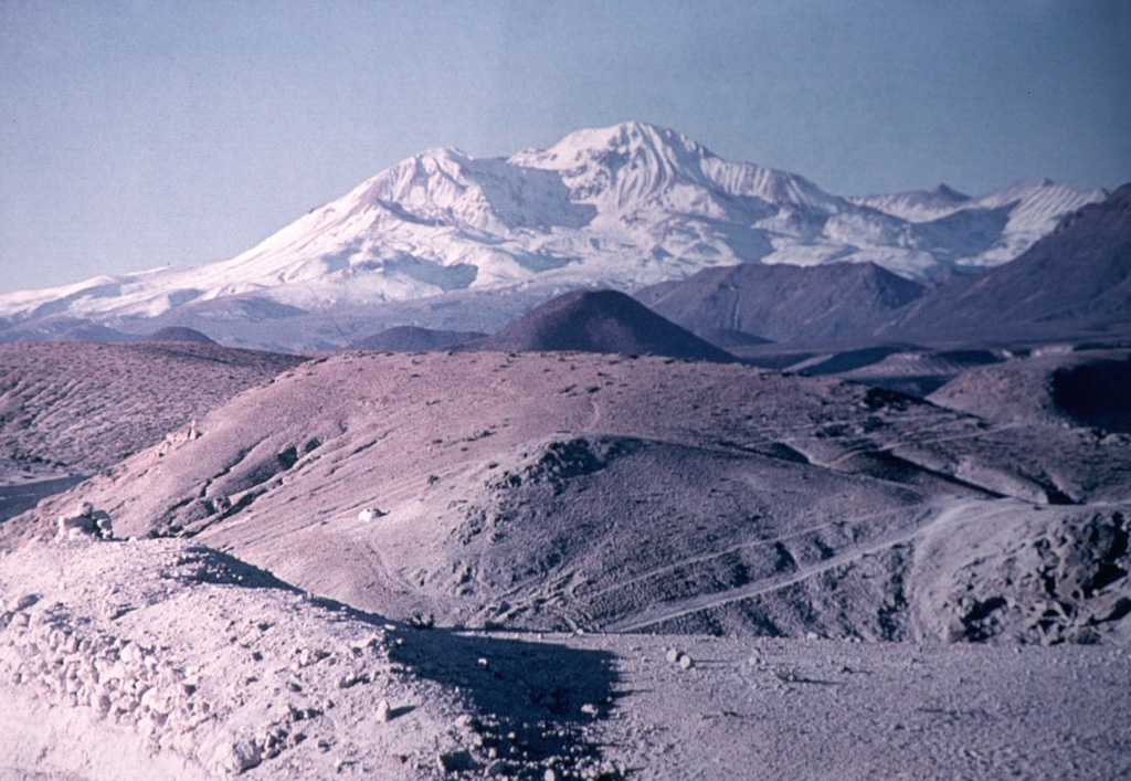 The snow-capped Taapaca (Nevados de Putre) volcanic complex rises to the north above rhyodacitic pyroclastic deposits in the Pampa del Muerto. Taapaca volcano rises NE of the town of Putre in northern Chile.  Putre is built on top of debris-avalanche deposits from Taapaca, which consists of a dacitic lava-dome complex.  The latest stage of activity during the Holocene produced the 5860-m-high summit lava dome complex (center horizon). Photo by Oscar González-Ferrán (University of Chile).