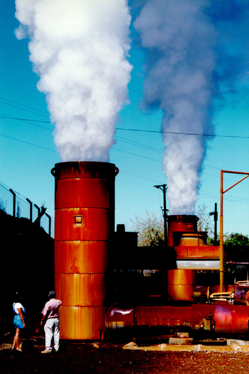 Steam pours from twin silencer towers at the Berlín 2 geothermal site.  Geothermal fluids can flow from a well at the speed of sound, which resembles that of a jet engine to the field crew.  The silencer drops the sound to a dull roar.  Two 25 MW power plants came on line at Berlín 2 in 1998 and 1999.  The two plants utilize both production and reinjection wells, and were expected (after operating and financial expenses) to save $56,000 in fuel costs per day. Photo courtesy of Comisión Ejecutiva Hidroeléctricia del Río Lempa (CEL), 1992.