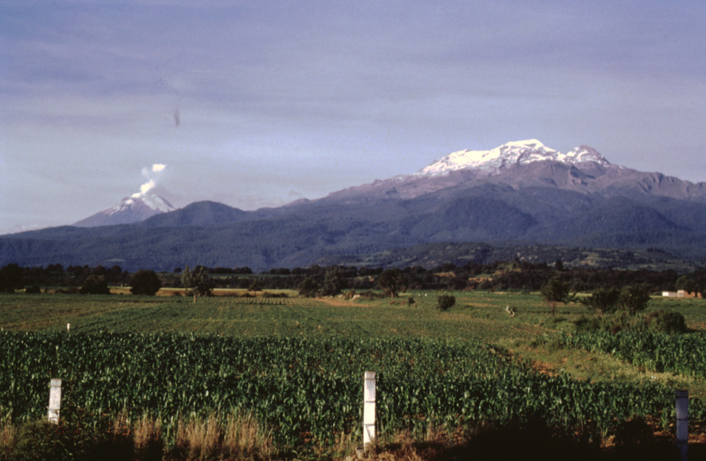 Iztaccíhuatl (right) and Popocatépetl (left) rise above farmlands at the eastern margin of the Puebla basin within Izta-Popocatépetl National Park. Andesite and dacite lava flows erupted from vents along the N-S-trending summit ridge of Iztaccíhuatl extend all the way to the base of the volcano. A small plume rises from the summit of Popocatépetl in this 1996 photo. Photo by José Macías, 1996 (Universidad Nacional Autónoma de México).