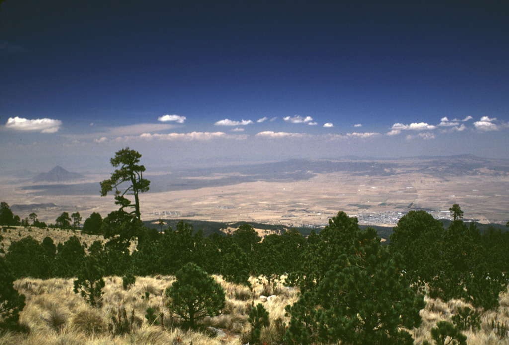 The light-colored area just below the right horizon is the floor of the 15 x 21 km Los Humeros caldera. It is seen here from the SE across the Serdán-Oriental basin from the flanks of Cofre de Perote volcano. Caldera formation during the mid-Pleistocene was followed by extrusion of voluminous lava flows during the late-Pleistocene or Holocene. These flows form the dark-colored area extending across the basin. The peak in the distance to the left is Cerro Pizarro. Photo by Lee Siebert, 1999 (Smithsonian Institution).