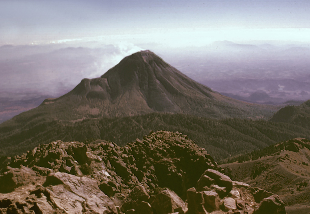 Colima volcano is seen here from the summit of neighboring Nevado de Colima. The forested ridge extending across the center of the photo is the rim of a large scarp formed by the collapse of the ancestral Colima edifice on the southern flank of Nevado. The modern Colima edifice grew within this scarp during the past few thousand years. The ridge on the NE (left) flank of Colima is El Volcancito, a lava dome that formed in 1869. Photo by Lee Siebert, 1998 (Smithsonian Institution).