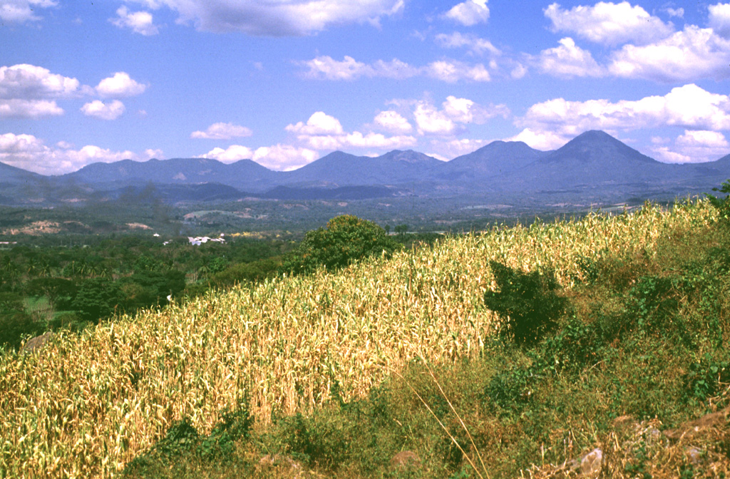 The Apaneca Range is seen here from the SE. This elongated group of Pleistocene and Holocene edifices lies in western El Salvador, between the Santa Ana complex and the Guatemala border. The 5-km-wide Pleistocene Concepción de Ataco caldera lies beyond the flat ridge to the left; to its right are Cerro Cachio, Cerro de las Rana, Cerro el Aguila, and conical Cerro los Naranjos. The Apaneca Range is the site of the major geothermal field of Ahuachapán. Photo by Lee Siebert, 1999 (Smithsonian Institution).