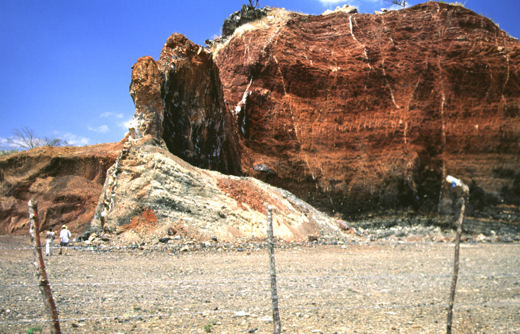 Volcanic dikes of varying orientations were emplaced into oxidized red scoria in a quarried scoria cone near the village of Agua Blanca. Several generations of thin dikes are visible (note people at the lower left for scale). The larger dike supporting the pinnacle near the center is oriented north-south, parallel to the direction of faults defining the Ipala graben. Photo by Giuseppina Kysar, 1999 (Smithsonian Institution).