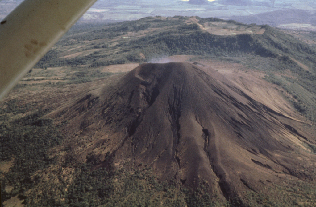 Telica's impressive summit crater is not visible in this aerial view from the west.  Erosional gullies scar the flanks of the unvegetated cone.  The broad ridge in the background is El Liston, an older part of the Telica volcanic complex.  An E-W-trending line of shallow craters extends from Telica to the broad summit of El Liston. Photo by Jaime Incer, 1991.