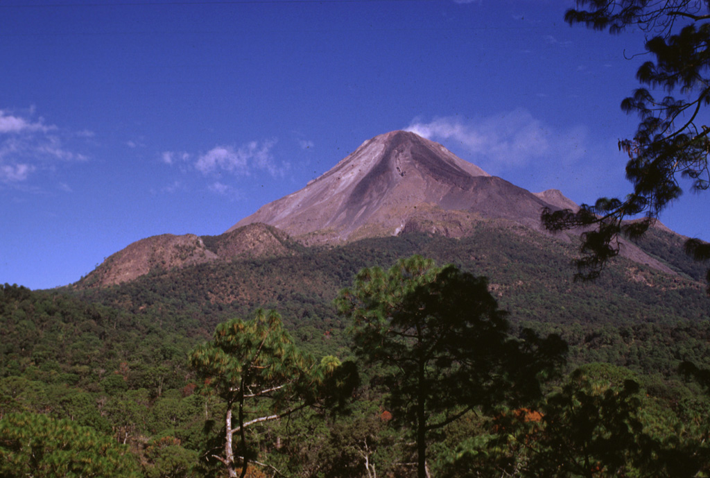 The two rounded peaks to the lower left are the Los Hijos del Volcán lava domes on Colima. These domes formed immediately south of the partially buried remnant of the SE caldera wall. The two domes and associated lava flows post-date caldera formation and mark the southernmost volcanic center of the Cantaro-Colima volcanic chain. Another flank lava dome, El Volcancito, forms the small ridge on the lower right flank, NE of the degassing summit. Photo by Lee Siebert, 1993 (Smithsonian Institution).