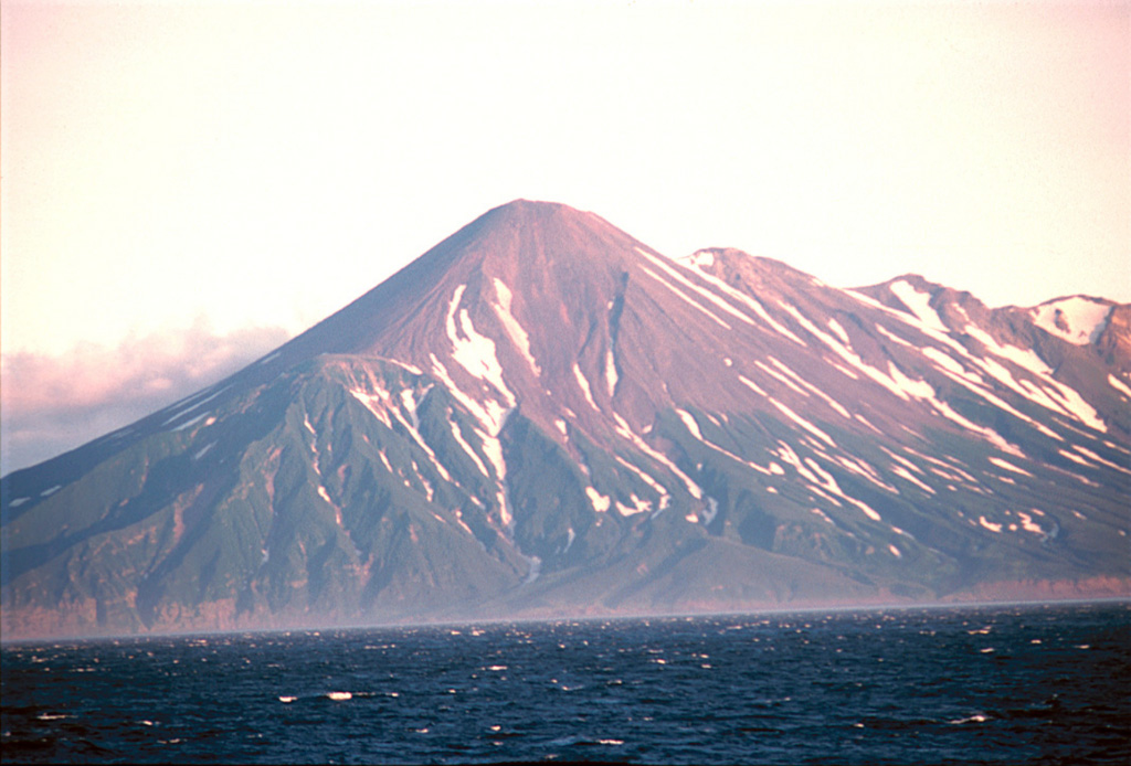 The NW slopes of Chikurachki, the highest volcano on Paramushir Island, rises above the Sea of Okhotsk. Oxidized scoria covering the upper part of the cone result in the distinctive red color. The Tatarinov group of six volcanic centers is located immediately to the south of Chikurachki, forming the ridge to the right. In contrast to the frequently active Chikurachki, the Tatarinov volcanoes are extensively eroded and have a more complex structure. Photo by Yoshihiro Ishizuka, 2000 (Hokkaido University).