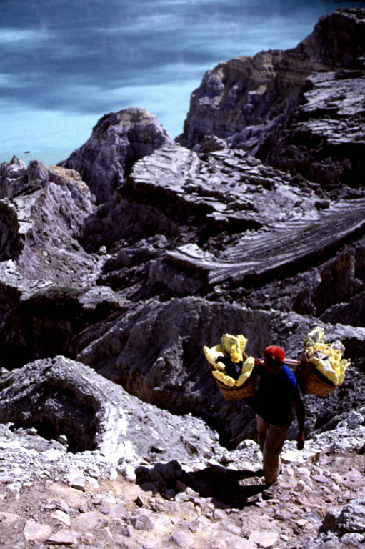 A sulfur miner carries baskets of elementally pure sulfur from a geothermal area near the shore of Kawah Ijen lake in eastern Java. Large blocks of sulfur are broken off and carried out of the crater in baskets before being transported to a sulfur mill on the SE flank of the Ijen caldera. The perspective in this view from near the SE crater lake rim looks down on the surface of the crater lake. Photo by Lee Siebert, 2000 (Smithsonian Institution).