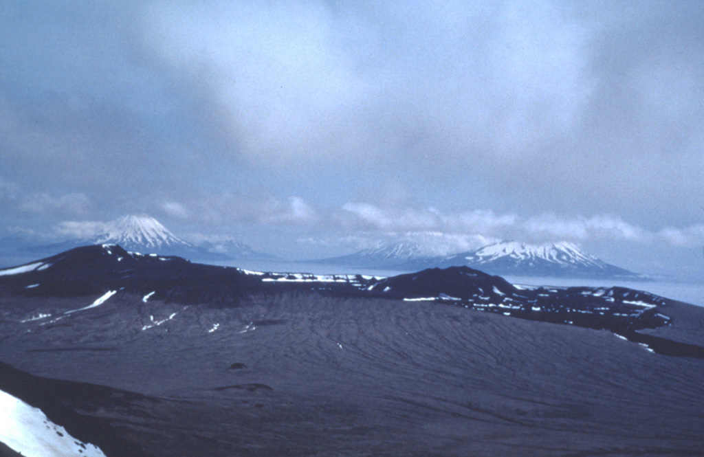 The summit caldera rim of Yunaska is seen from an older volcanic center on the western side of the island. The younger eastern complex contains two nested calderas. A young lava flow to the far right descended through a notch in the caldera rim. The peaks in the background are (left-to-right) of Carlisle, Cleveland, and Herbert volcanoes. Photo by Jim Meyers, 1992 (University of Wyoming, courtesy of Alaska Volcano Observatory).