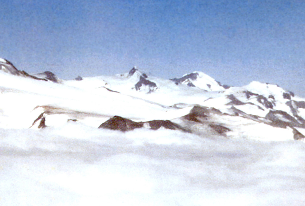The rounded peak of Mount Steller is visible on the horizon at right-center beyond the slopes of Denison; the summit ridge of Snowy volcano forms the far-left skyline in this long-distance NE-looking view. The slopes of Mount Denison are seen on the right half of the photo, between the pointed summit in the center and the flatter peak to the far right, between which a glacier descends towards the middle of the image.  Photo courtesy of Alaska Volcano Observatory, U.S. Geological Survey, 1997.