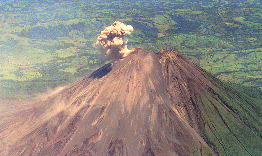 An ash plume rises from the active summit crater of Arenal in this April 1998 view from the south. A small saddle separates the unvegetated post-1968 cone from the pre-1968 cone (right). During April 1998 lava flows traveling down the northern-to-western flanks, and blocks on the NW Crater C wall, broke loose and tiggered small avalanches. This relatively minor activity preceded a larger eruption that produced pyroclastic flows on 5 May.  Photo by Federico Chavarria Kopper, 1998.