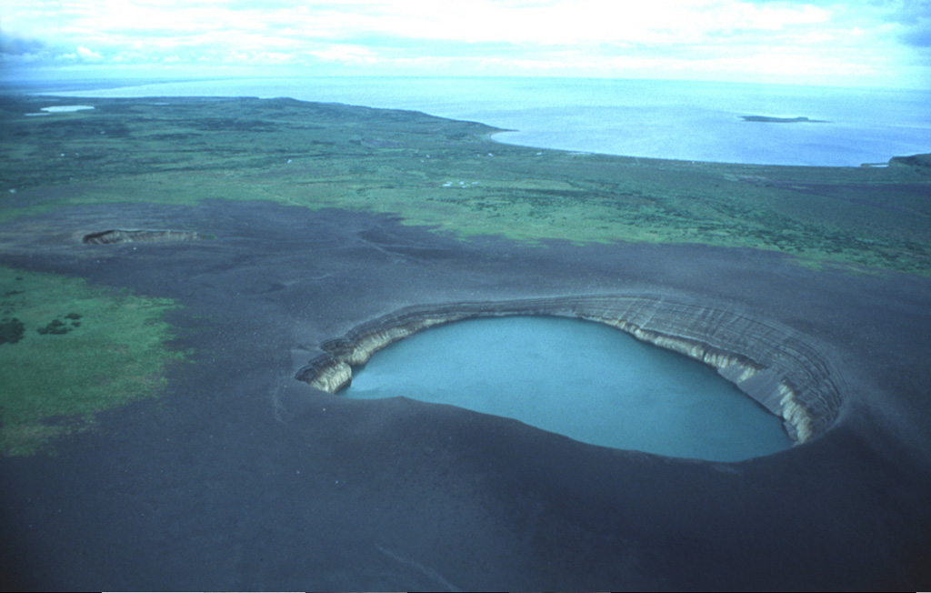 This lake-filled maar and another small crater (left) are the Ukinrek Maars, which formed during an eruption in 1977 in a lowland area of the Alaska Peninsula west of the main volcanic chain. The contact between lighter and darker material in the walls of the lake-filled eastern maar marks the original pre-eruption surface, which is now buried by a dark apron of ejecta produced during the eruption. Photo by Christina Neal, 1993 (Alaska Volcano Observatory, U.S. Geological Survey).