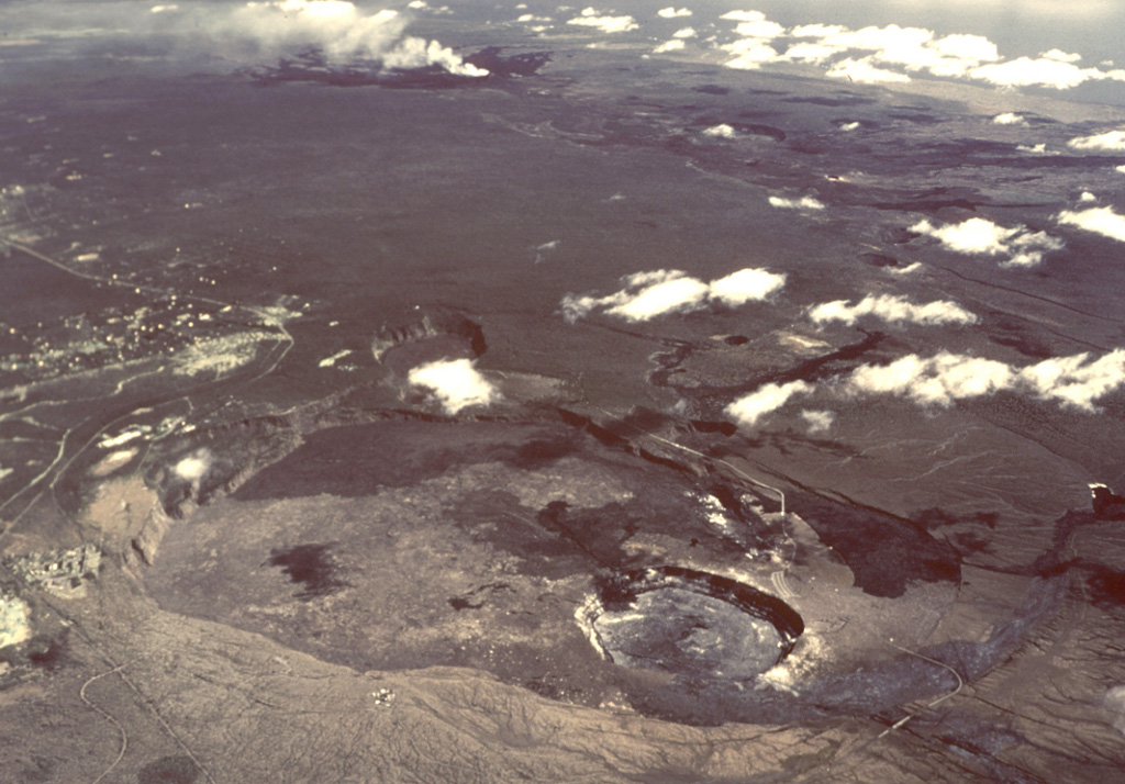 An aerial view in 1985 looks from Kilauea caldera in the foreground down the East Rift Zone to the  Puʻuʻōʻō vent producing a gas plume in the background. The Hawaiian Volcano Observatory lies at the lower left on the western rim of Kilauea's 5-km-wide summit caldera with the inner Halema’uma’u crater to the lower right. Lava flows cover the floor of the summit caldera and can be seen extending downslope from multiple vents along the rift zone, including the dark-colored most recent flows erupted beginning in 1983 from the Kupaianaha vent. Photo by Jim Griggs, 1985 (U.S. Geological Survey).