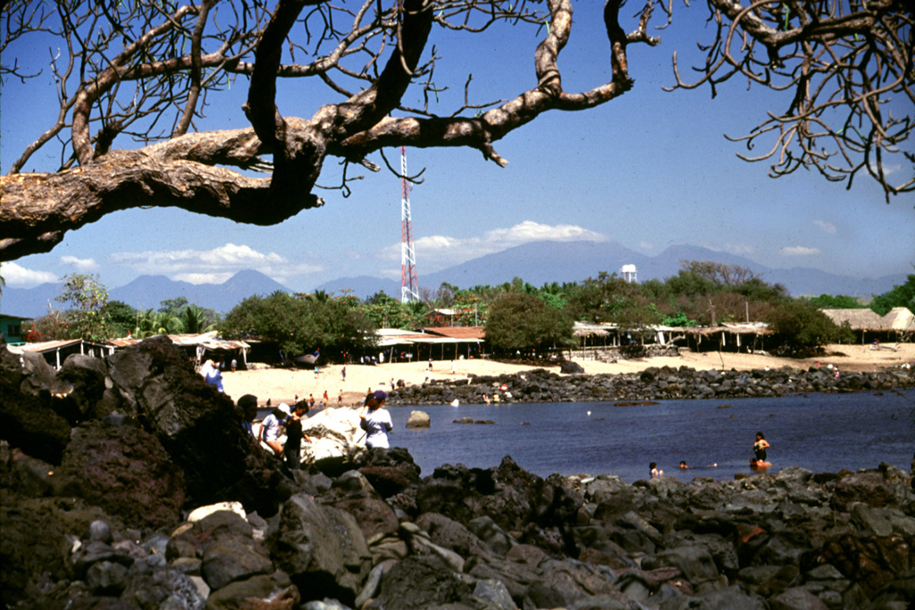 The massive Acajutla debris avalanche swept into the sea, forming a peninsula that extended the shoreline about 7 km. The rocks in the foreground and across the bay at Playa Los Cobanos are the farthest subaerial extent of the avalanche, 41 km from its source at Santa Ana volcano on the horizon to the right. Hummocks are exposed to the coastline and are visible well offshore on bathymetric surveys, which suggest that the avalanche deposit has a significant submarine component. Photo by Lee Siebert, 2002 (Smithsonian Institution).