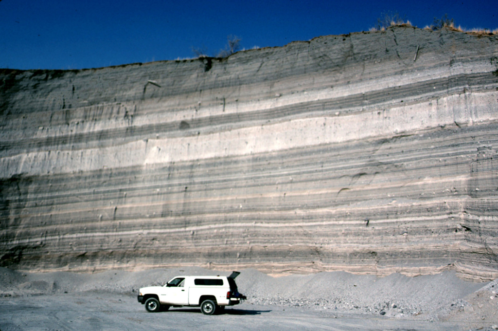 The walls of a quarry on the SW flank of Hoya Estrada maar close to the city of Valle de Santiago show a spectacular sequence of deposits from the maar-forming eruptions.  Most of the outcrop consists of gray-colored dominantly planar pyroclastic surge beds. The three prominent light-colored layers are ashfall deposits. The largest ash layer is about 2 m thick just above the middle of the outcrop in this view looking towards the vent. Photo by Jim Luhr, 2002 (Smithsonian Institution).