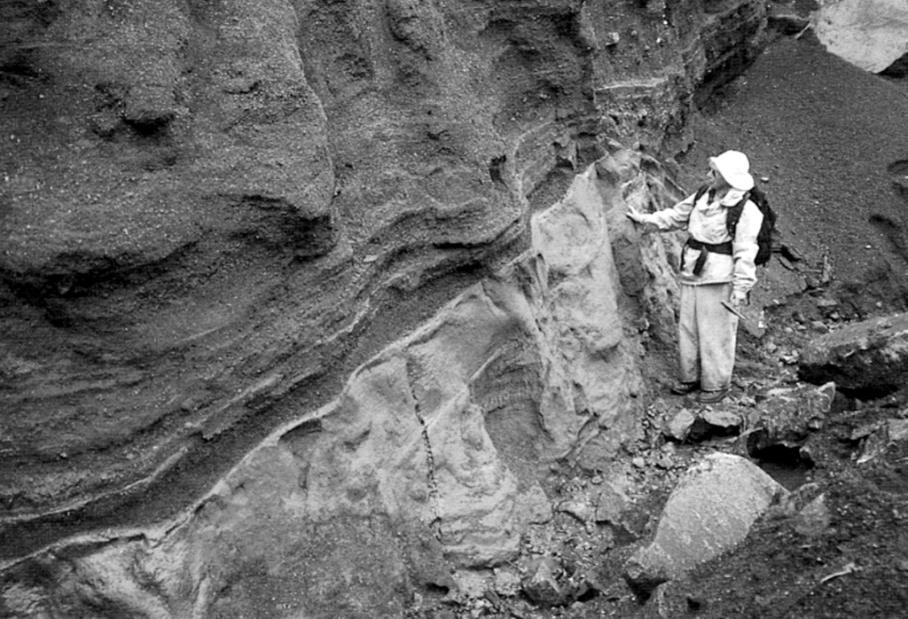 A geologist observes the contact between a basaltic Plinian fallout lapilli unit overlying marine clay. This deposit is part of thick beds of basaltic Holocene tephra originating from an unknown Quaternary volcanic center found near Crow Lagoon, north of Prince Rupert near the southern tip of the Alaskan panhandle. Ballistically emplaced bombs imply a nearby source. The tephra beds are located along the south side of the Khutzeymateen Inlet, about 40 km N of Prince Rupert. Photo by Jack Souther (Geological Survey of Canada, courtesy of Cathie Hickson).