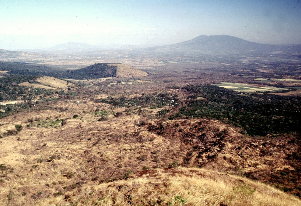 The Teixcal lava flow is the brownish area extending diagonally from the lower left towards San Salvador volcano on the right horizon. It originated during an eruption in 1722 from the base of San Marcelino scoria cone, where this photo was taken. The lava is noted for its disequilibrium textures, where large orthopyroxene crystals are surrounded by olivine reaction rims. The half-forested, half-vegetated cone near the center is Cerro Alto, a scoria cone of Coatepeque caldera (out of view to the left). Photo by Lee Siebert, 2002 (Smithsonian Institution).