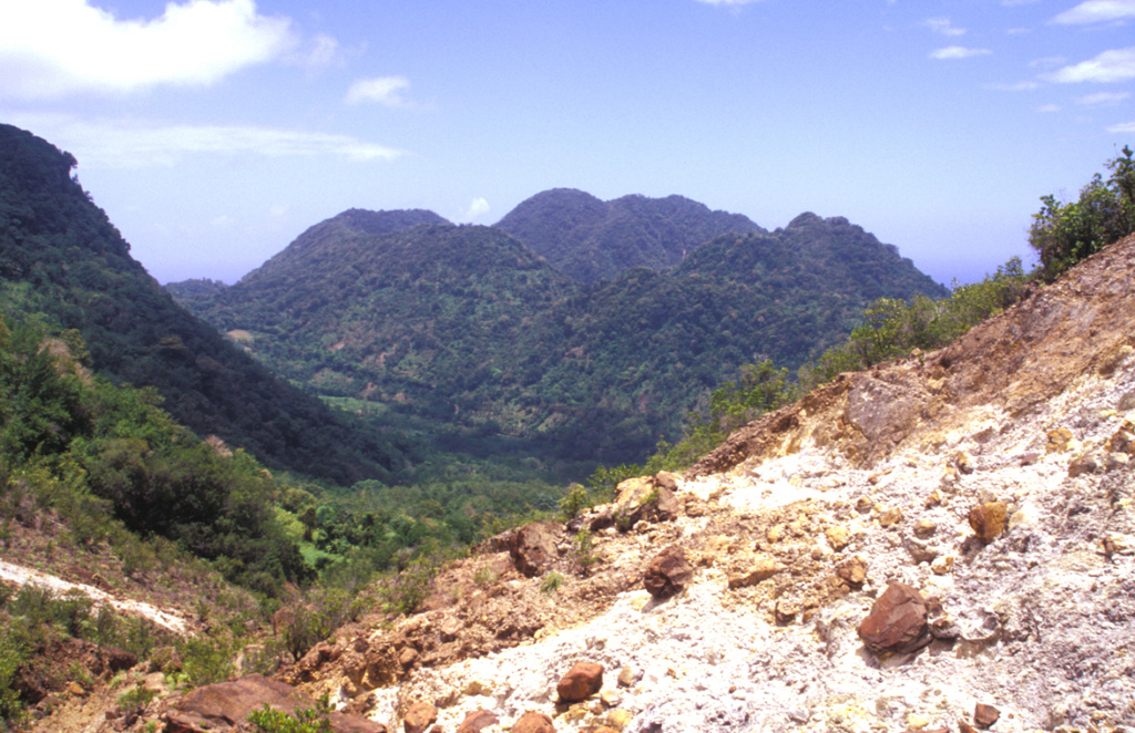 The youngest cluster of lava domes of the Morne Plat Pays volcanic complex, Morne Rouge, Crabier, and Morne Patates (left to right) is seen here from the north at the upper Sulfur Springs thermal area.  The cliff in the shadow at the left is the west-facing wall of the arcuate Morne Plat Pays caldera, which formed about 39,000 years ago in association with the eruption of the Grand Bay Ignimbrite and a lateral flank collapse to the west. Photo by Paul Kimberly, 2002 (Smithsonian Institution).