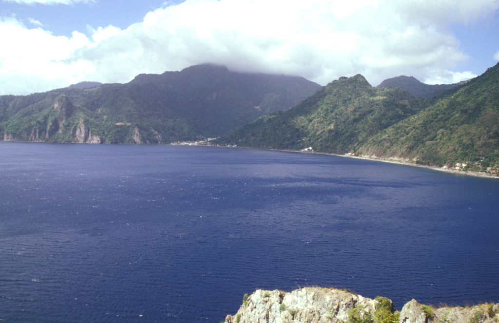 The promontory at Scotts Head provides a vantage point overlooking the partially submerged Plat Pays caldera, widely breached to the west at Soufrière Bay.  The caldera, which partially truncated Morne Plat Pays stratovolcano (in the clouds on the center horizon) formed about 39,000 years ago.  Its margins extend from north of Sorcière lava dome (far left) to near Scotts Head.  Morne Patates, the dome along the coast at the right-center, is the youngest post-caldera lava dome and was active until at least about 700 years ago. Photo by Paul Kimberly, 2002 (Smithsonian Institution).