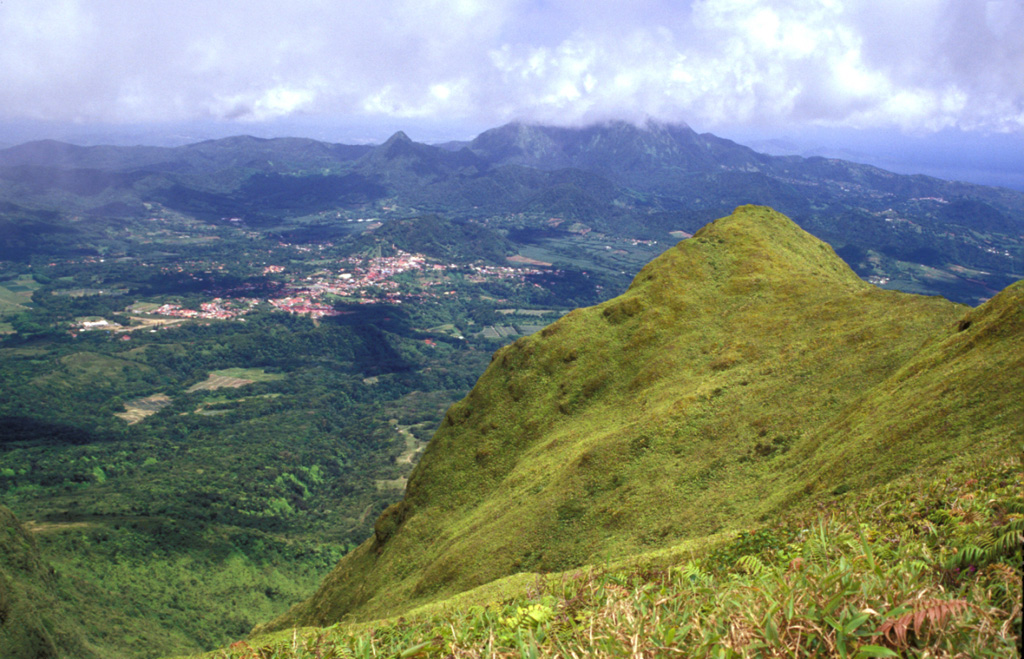The steep-sided grassy knob in the right foreground is part of the Aileron lava dome, which formed during an eruption about 9700 years ago.  This view looks to the SE towards the town of Morne Rouge (left-center), which was devastated by pyroclastic flows during the 1902 eruption.  The Pleistocene Piton du Carbet volcano lies in the clouds on the right-center horizon. Photo by Lee Siebert, 2002 (Smithsonian Institution).