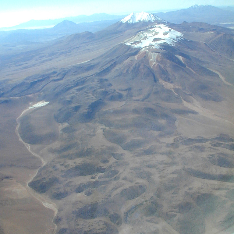 The Olca-Paruma volcanic complex, seen here from the west, forms a 15-km-long E-W ridge forming the border between Chile and Bolivia and is comprised of several stratovolcanoes with Holocene lava flows.  Volcán Olca lies near the western end of the complex.  It is flanked to the east by Volcán Paruma, which is immediately west of the higher pre-Holocene Cerro Paruma volcano, the conical peak in the background.  Volcán Paruma has been the source of conspicuous fresh lava flows and has displayed persistent fumarolic activity in recent years.    Photo by José Naranjo, 2001 (Servico Nacional de Geologica y Mineria).