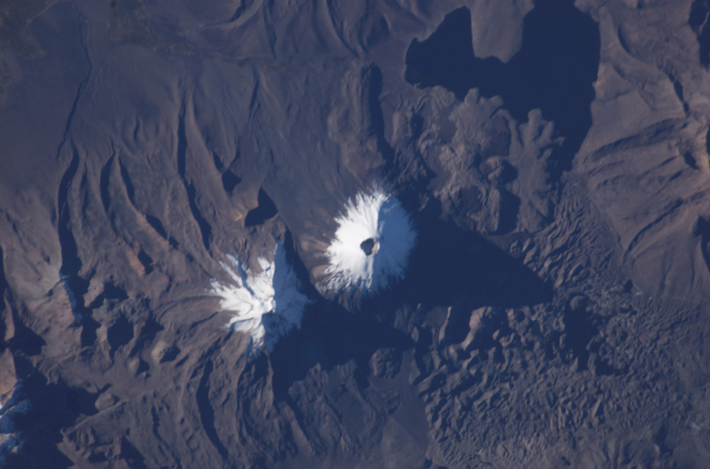 The two snow-capped volcanoes of the Nevados de Payachata volcanic group dominate this NASA International Space Station image (with north to the bottom).  A prominent summit crater tops symmetrical Parinacota volcano, constructed to the SW of its eroded Pleistocene twin, Pomerape volcano.  Silicic lava flows from Parinacota form lobes extending into Laguna Chungará, which was formed when a major debris avalanche from Parinacota blocked drainages.  The hummocky debris-avalanche deposit covers much of the lower right part of the image. NASA International Space Station image ISS009-E-6837, 2004 (http://eol.jsc.nasa.gov/).