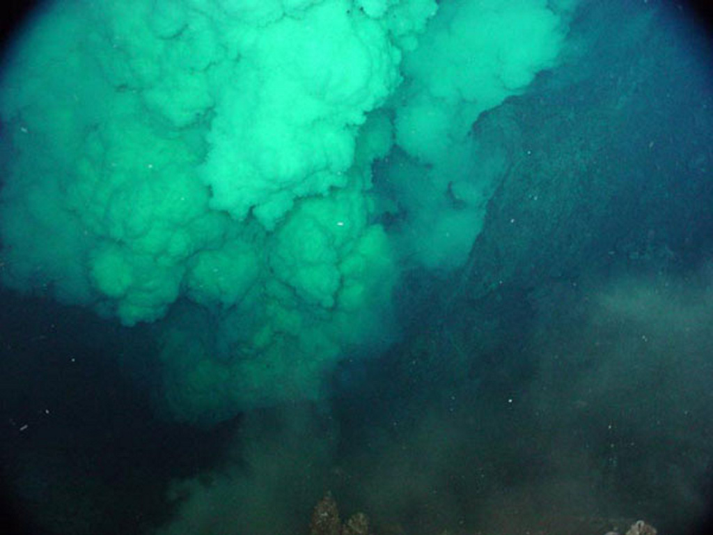 A dense, particle-laden plume rises above the NW Rota-1 Brimstone Pit vent in this 30 March 2004 photo taken from a remotely operated vehicle (ROV) during a NOAA bathymetric survey of the Mariana arc. The vent intermittently ejected a plume several hundred meters high containing ash and molten sulfur droplets that adhered to the surface of the ROV. At other times during the exploration venting slowed or ceased, allowing views of the funnel-shaped vent that was about 20 m wide and 12 m deep. NOAA image, 2004 (courtesy of Bill Chadwick; http://oceanexplorer.noaa.gov/explorations/04fire/logs/march30/march30.html).