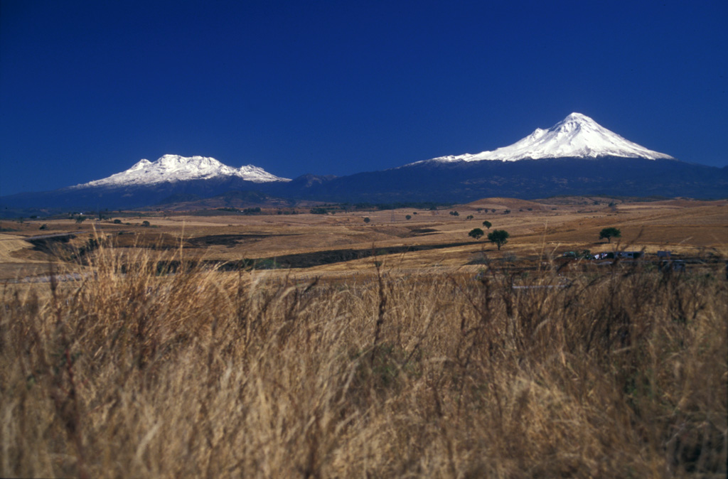 Iztaccíhuatl (left) and Popocatepetl (right), two of the highest volcanoes in Mexico, are seen from the SW on a rare smog-free day. The morphology of Iztaccíhuatl is due to multiple vents across the summit region . Fresh snowfall at Popocatepetl covers periodic ashfall deposits.  Photo by Lee Siebert, 2004 (Smithsonian Institution).