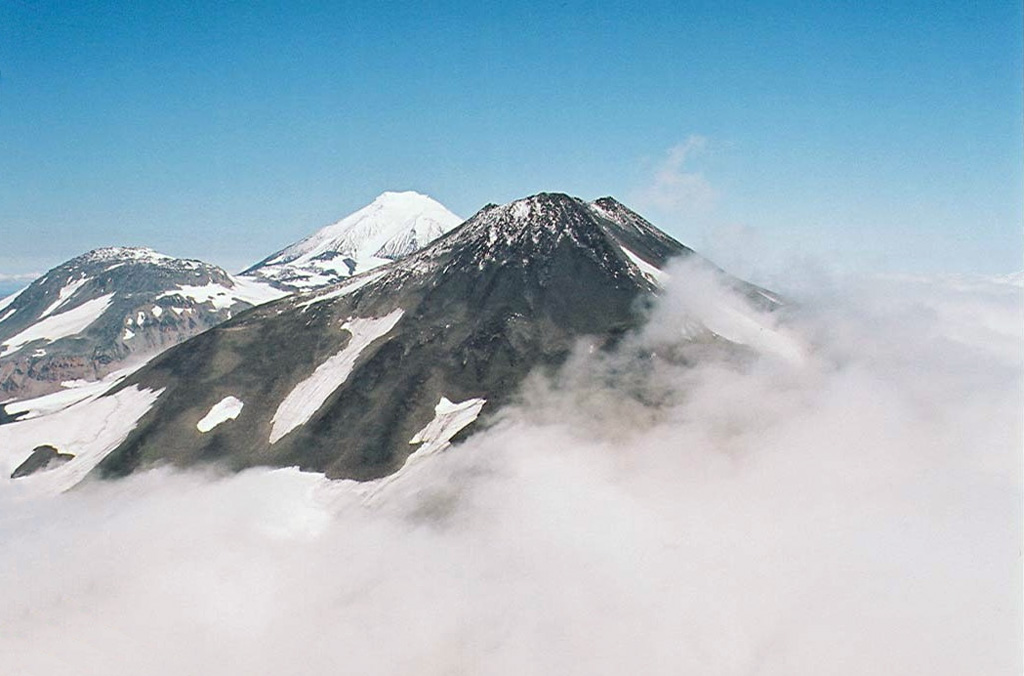 Three volcanoes in the northern Sredinny Range rise above the clouds in this helicopter view from the south. Spokoiny is in the foreground, with snow-covered Ostry in the background. The rounded lower peak on the west (left) flank of Ostry was active during the Holocene. Copyrighted photo by Maria Pevzner, 2005 (Russian Academy of Sciences, Moscow).