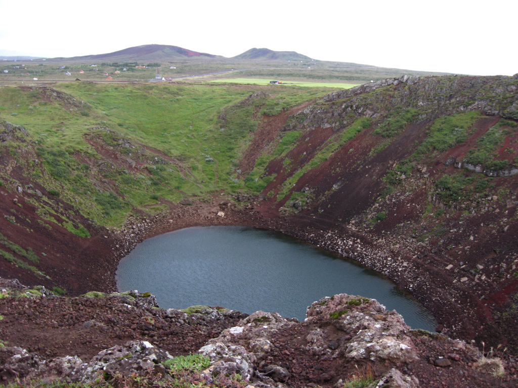 A small pond about 60 x 100 m fills the bottom of the Kerid crater at the northern end of the Tjarnarhólar crater row in the Grímsnes volcanic system. The crater is elliptical in shape, 180 x 280 m. The Seyðishólar and Kerhóll cinder cones, visible in the background to the north, were active about 9,500 and 7,050 years ago respectively. Seyðishólar is the only Holocene eruption recognized to have had tephra dispersal outside of the volcanic field. Photo by Lee Siebert, 2008 (Smithsonian Institution).