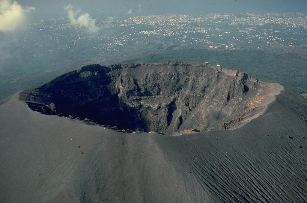 The summit crater of Vesuvius volcano exposes the layered lava flows and ash beds that have constructed the cone.  Ash deposits from the 1944 eruption mantle the rim and outer flanks of the 600-m-wide, 300-m-deep crater.  The Bay of Naples and numerous cities along the coastal plain that have been affected by historical eruptions of Vesuvius can be seen at the top of the photo, taken from the NE. Copyrighted photo by Katia and Maurice Krafft, 1982.