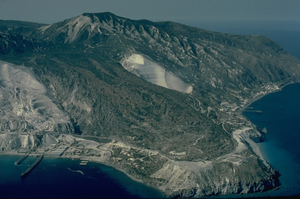 The Rocche Rosse obsidian flow in the foreground forms a peninsula at the NE tip of Lipari Island.  It, along with the Forgia Vecchia obsidian flow to the south, was emplaced during the last eruption of Lipari in 729 CE.  The lava flows were emplaced following explosive eruptions that ejected large amounts of pumice.  The light-colored areas at the center and left are large-scale pumice quarries.  Copyrighted photo by Katia and Maurice Krafft.