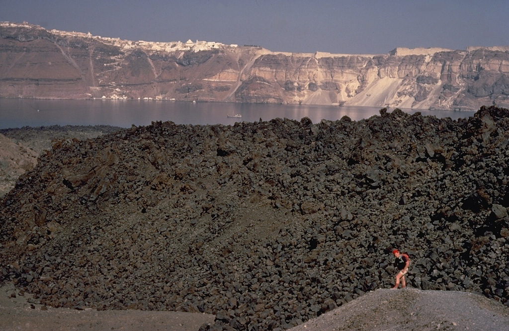 The small, blocky lava dome in the foreground is the product of an eruption of Santorini volcano during 1939-1941.  This eruption, smaller than previous post-Minoan eruptions of Santorini, began with phreatic explosions, and concluded with the effusion of the dome and small lava flows.  French volcanologist Maurice Krafft provides scale in the foreground, and the steep walls of Santorini's largely submerged caldera, capped by the whitewashed village of Firá, rise to the NE across the caldera bay. Copyrighted photo by Katia and Maurice Krafft, 1970.