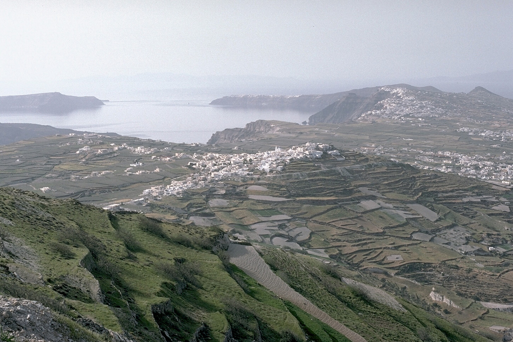 The outer flanks of the Santorini caldera, mantled by deposits of the 3,500-year-old Minoan eruption, provide a setting for croplands and island villages. This view looks NW from Mt. Profitis Ilias, a limestone peak forming the high point of the island of Thera, shows the northern half of the 7.5 x 11 km caldera. The northern rim drops below sea level, leaving a channel between the tip of Thera and the island of Therasia to the left. Photo by Lee Siebert, 1994 (Smithsonian Institution).