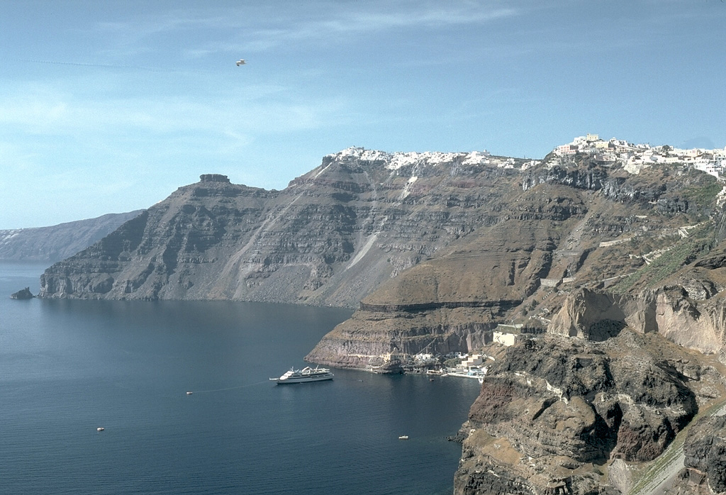The steep inner walls of Santorini drop steeply into the caldera bay. Pyroclastic flow deposits from four caldera-forming eruptions dating back to 100,000 years ago are exposed in the caldera walls in this N-looking view. The youngest caldera was formed about 3,500 years ago during the Minoan eruption of Santorini. The flat-topped peak on the left skyline is Skaros, a remnant of a shield volcano constructed within a previous caldera. Photo by Lee Siebert, 1994 (Smithsonian Institution).